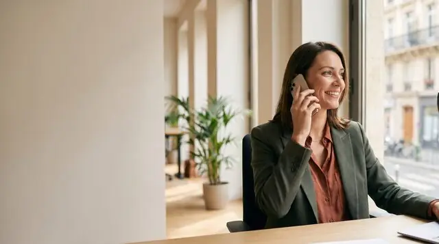 AI image: A smiling woman talks on a smartphone at a desk by a window – phone calls at work.